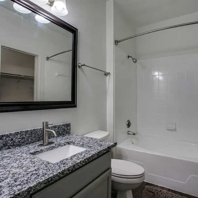 A modern bathroom featuring a gray granite countertop with a sink, a large mirror, and a bathtub with a shower. The walls are painted white, and the floor is adorned with a decorative mat.