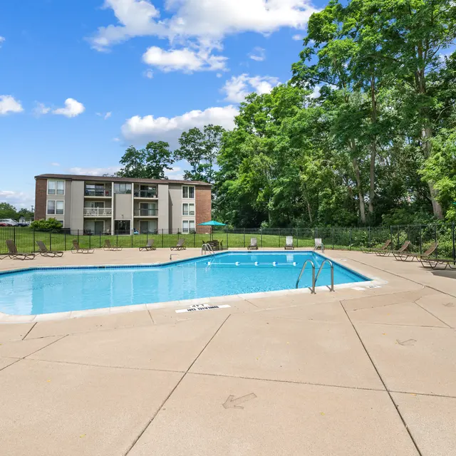 A clear swimming pool surrounded by a paved patio, lounge chairs, and umbrellas, with a residential building in the background and greenery on the sides.