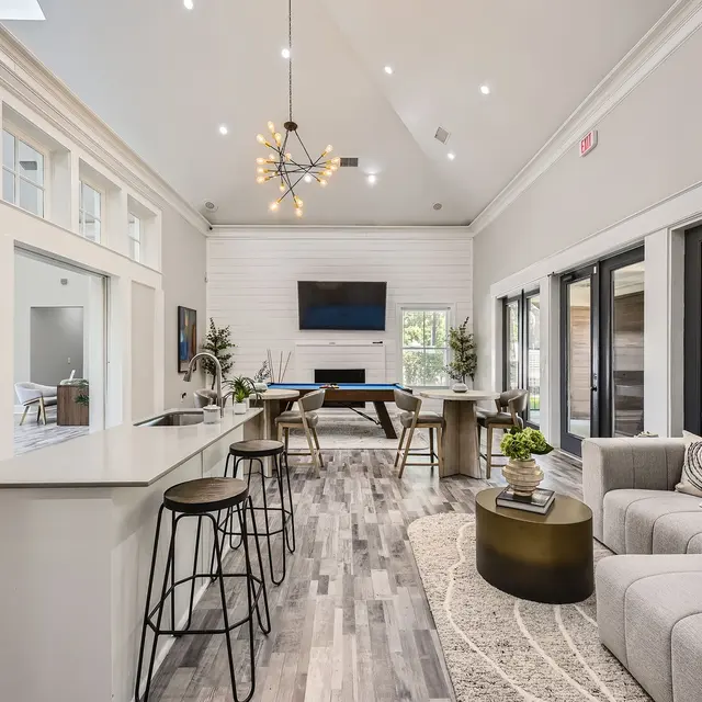 A spacious, modern living area with a high ceiling, illuminated by a stylish chandelier. The room features a large sectional sofa, a round coffee table, a dining area with wooden tables, and large windows letting in natural light. A flat-screen TV is mounted on the wall, and there are bar stools along the kitchen counter.