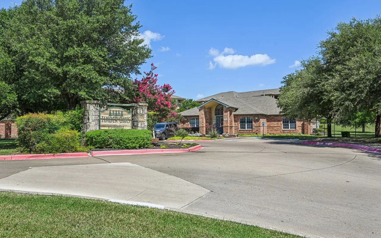 Entrance to a residential apartment complex with a stone sign and landscaping, under a clear blue sky.