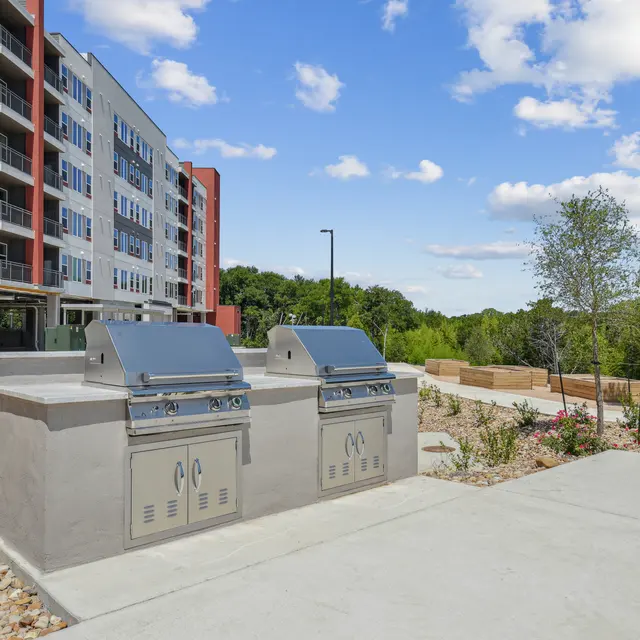Two stainless steel outdoor grills in a landscaped area with an apartment building in the background under a blue sky with fluffy clouds.