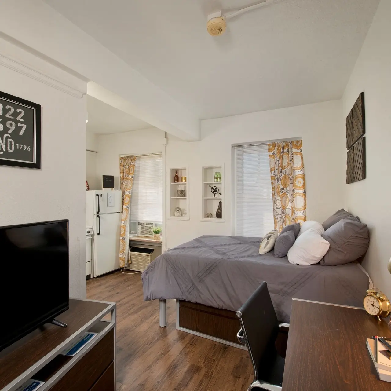 A cozy studio apartment featuring a bed with gray bedding, a small desk with a lamp and stationery, a television stand, and a kitchenette area with a refrigerator in the background. The windows are adorned with yellow patterned curtains.
