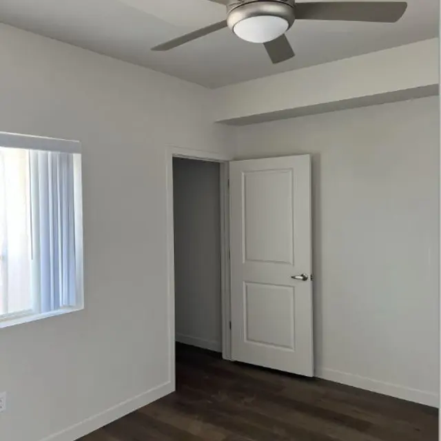 A modern, minimalistic room featuring a ceiling fan, a light-colored wall, and a window with blinds.