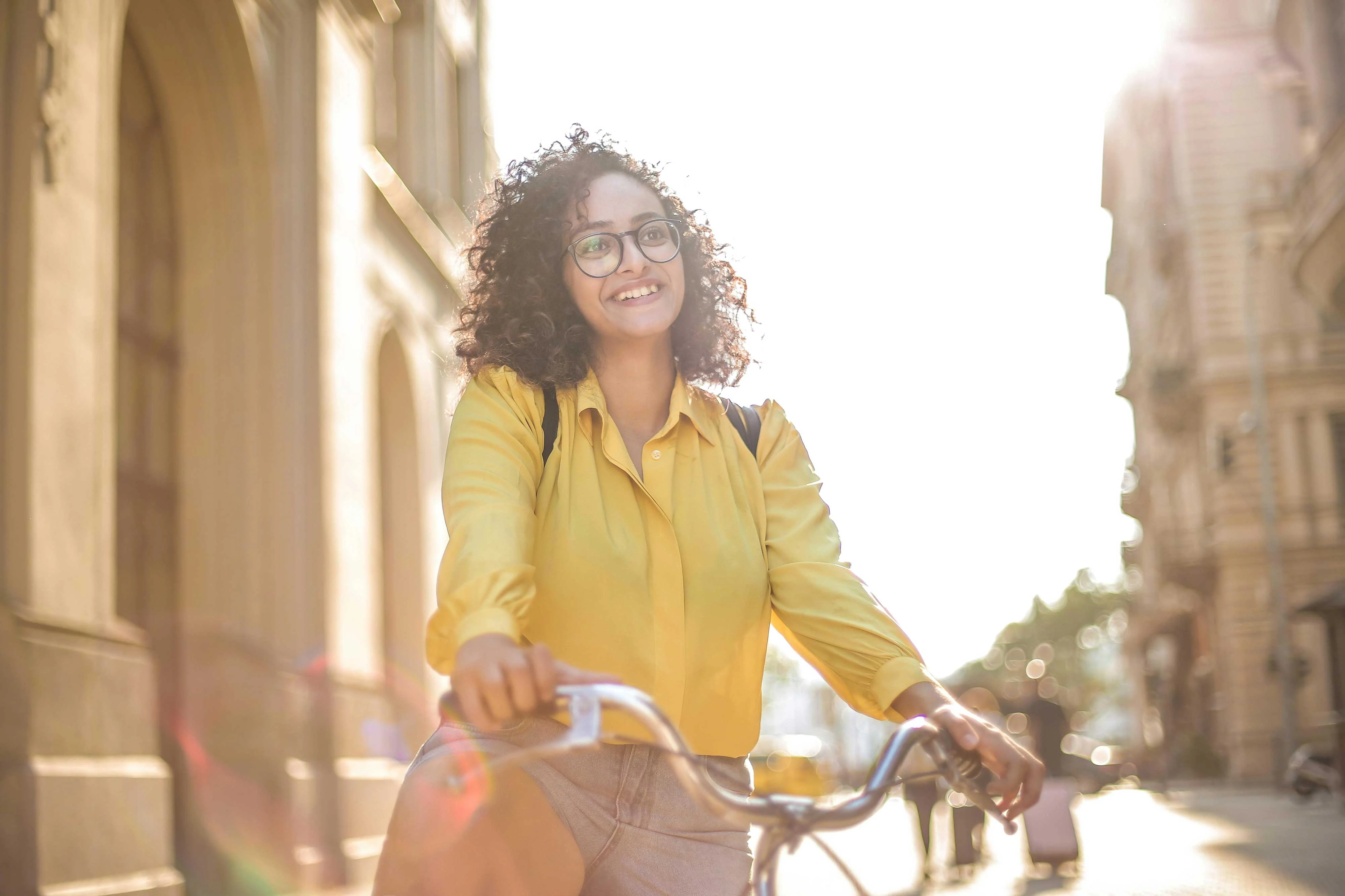 A young woman with curly hair and glasses smiles while riding a bicycle in a sunlit street.