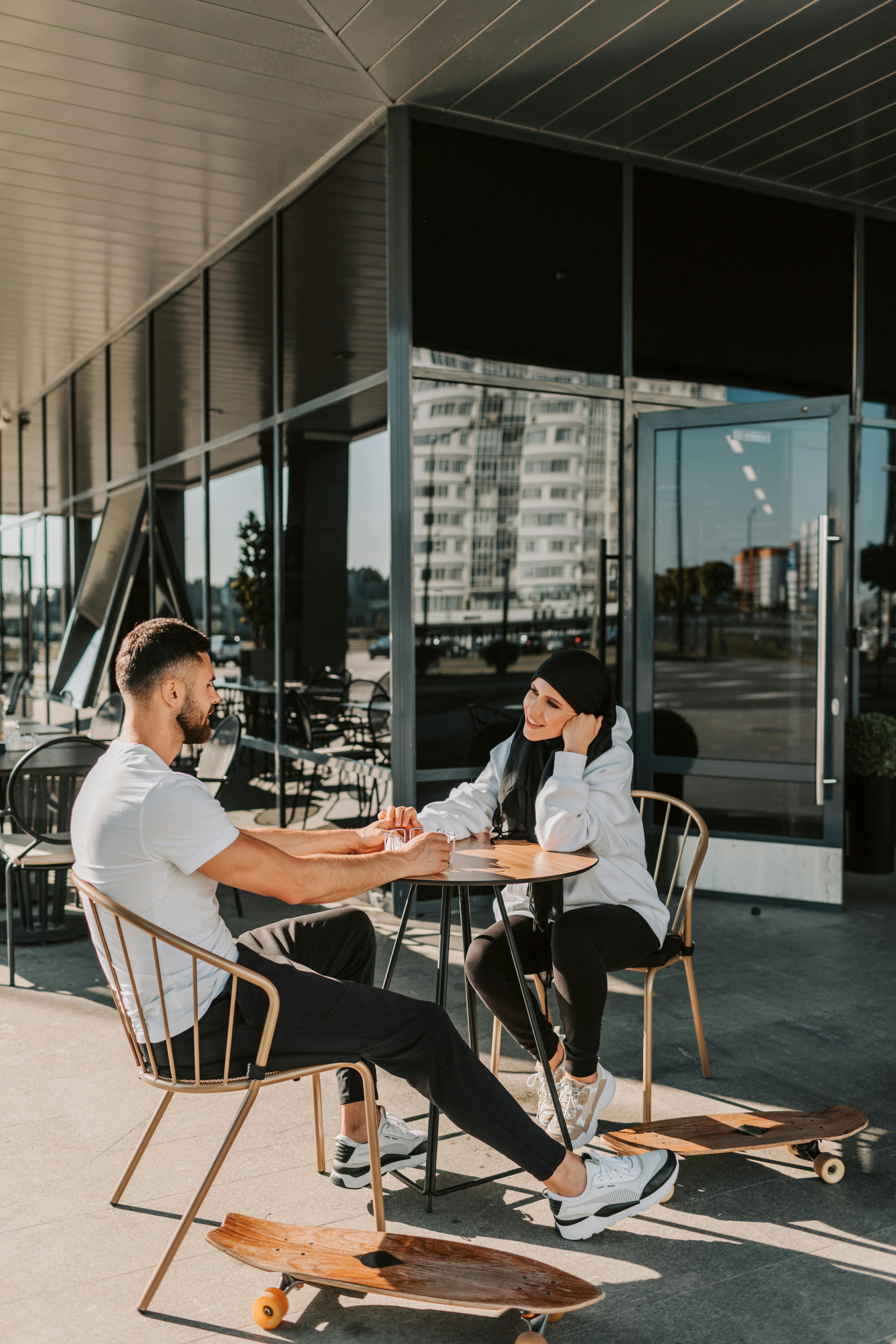 A couple sitting at a café table holding hands. The man is in a white t-shirt and black pants, while the woman, wearing a hijab and a jacket, looks at him with a smile. Two skateboards are placed on the ground near their chairs.