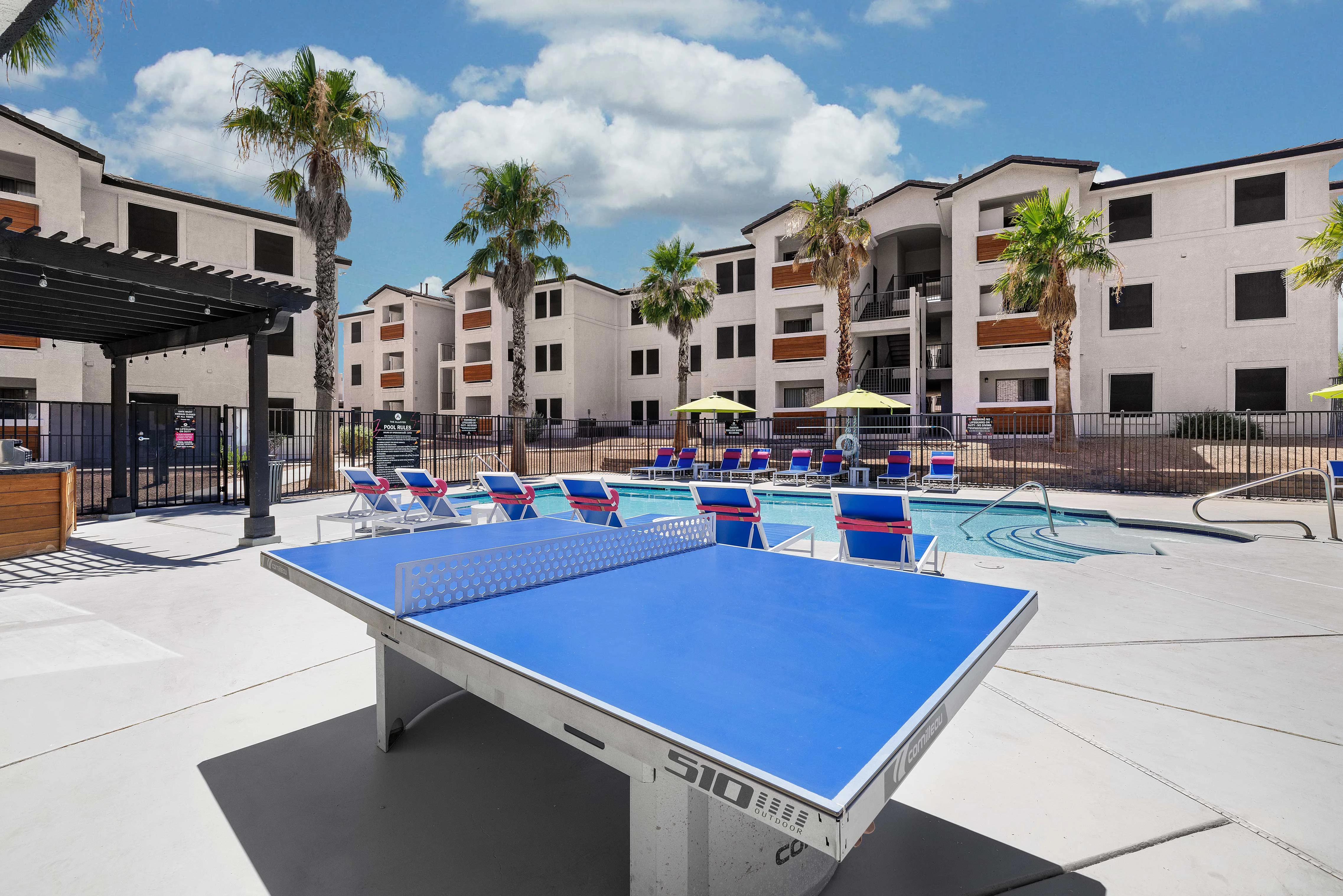 A bright, sunny apartment complex pool area featuring a blue ping pong table in the foreground, lounge chairs, and palm trees.