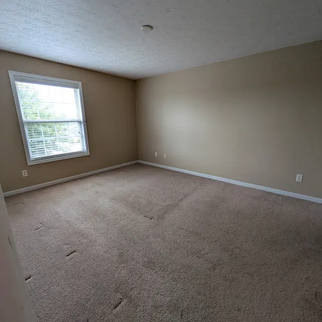 A vacant room with light brown walls and carpet, featuring a window with white blinds. The room has minimal furnishings and is well-lit.