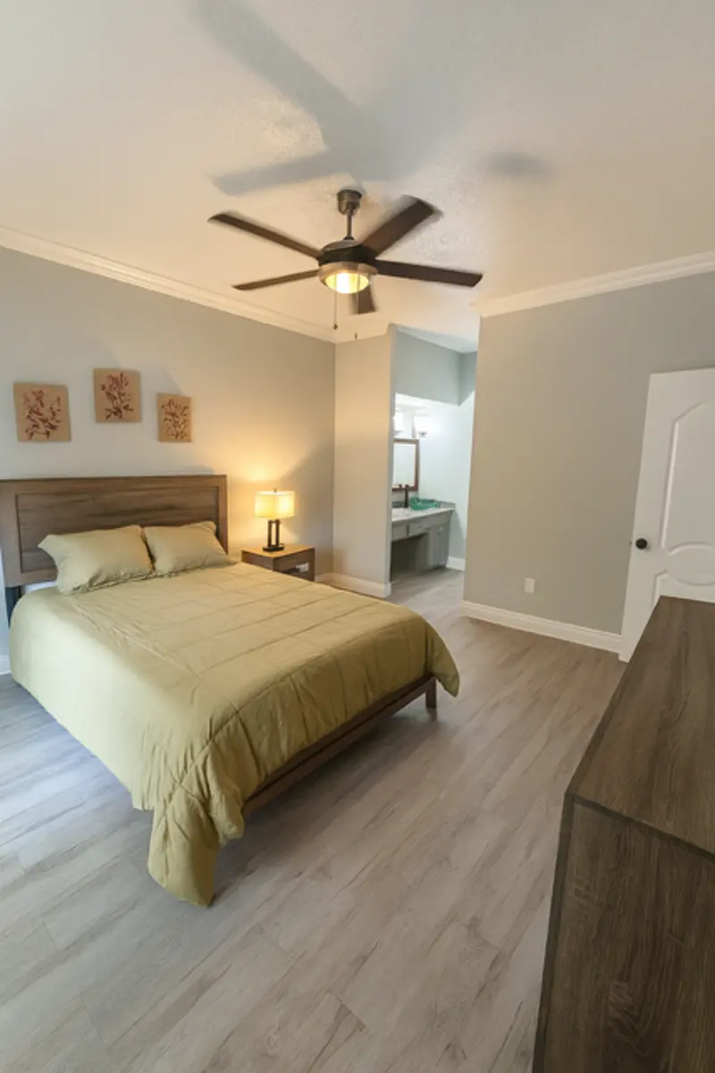 A modern bedroom featuring a light green bedspread, wooden furniture, and a ceiling fan. A lamp and decorative wall art are also present, with a separate area visible in the background.