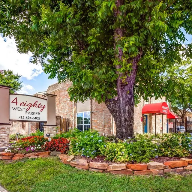 Exterior view of a building with a large sign reading '4 eighty WEST PARKER'. The area features landscaped gardens with flowers and shrubs, a large tree, and a sidewalk. A red awning is visible in the background.