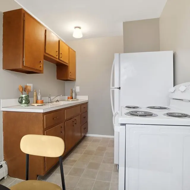 A small modern kitchen featuring wooden cabinets, a white refrigerator, and a white stove. The countertop is clear with some kitchenware and decorative items. A round table with a black top and yellow chair are visible.