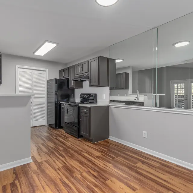 A modern kitchen featuring dark cabinetry, black appliances, and a reflective wall mirror. The kitchen has a wooden floor and a bar area.
