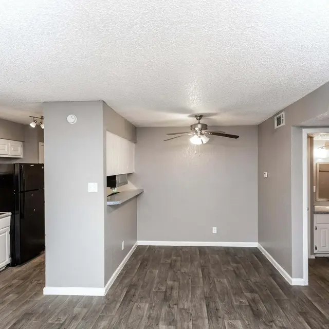 A contemporary apartment interior featuring an open layout with gray walls and wood-like flooring. The kitchen area is on the left, with white cabinets and black appliances. A ceiling fan is visible in the center, and there's a bathroom doorway on the right.
