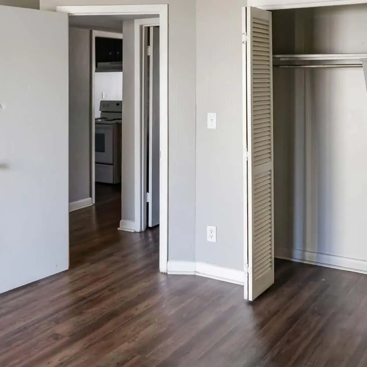 Haltere at Reserve - An empty room with gray walls and a wooden floor. There is a closet with sliding doors on the right, and a doorway leading to another room in the background. The room is well-lit, suggesting a bright atmosphere.