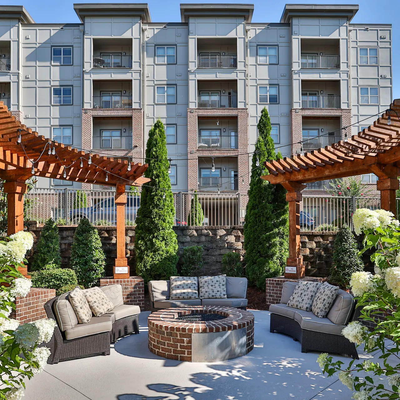Outdoor Lounge Area at Apartment Complex Outdoor lounge area with circular seating and a fire pit surrounded by greenery, overlooking a multi-story apartment building.