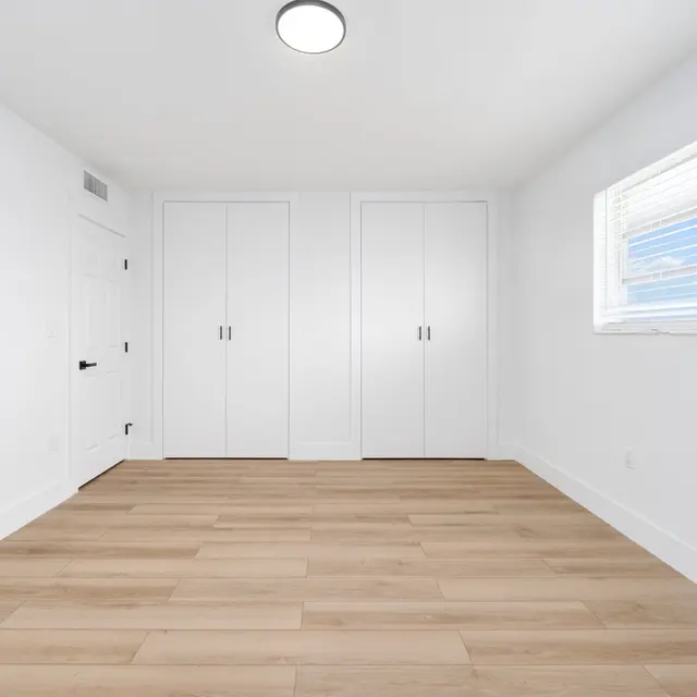 A minimalist bedroom with light wood flooring, white walls, and a bright window. There are two closets with white doors on one wall and a simple ceiling light fixture.