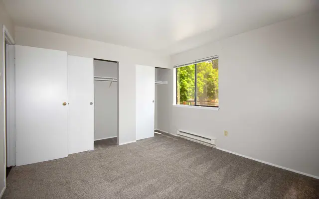 A spacious, empty bedroom featuring beige carpet, white walls, and light coming from a window. There are two white closet doors on the left side and a window to the right, showing greenery outside. No furniture is present in the room.
