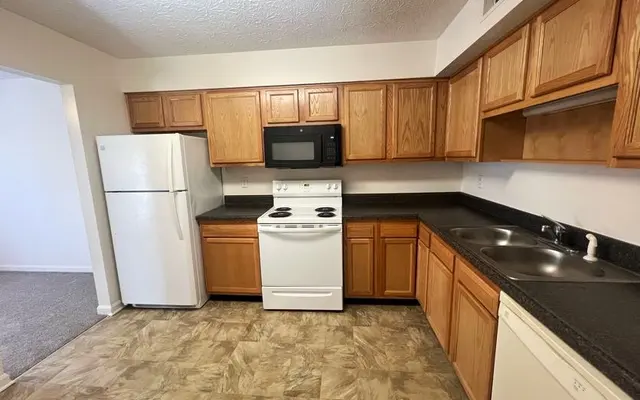 Modern Kitchen with Wood Cabinets A spacious kitchen featuring wooden cabinets, black countertops, a white refrigerator, a white stove, and a black microwave. The flooring is patterned, and there's a view into another room with carpet.