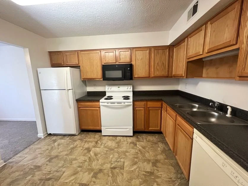 Modern Kitchen with Wood Cabinets A spacious kitchen featuring wooden cabinets, black countertops, a white refrigerator, a white stove, and a black microwave. The flooring is patterned, and there's a view into another room with carpet.