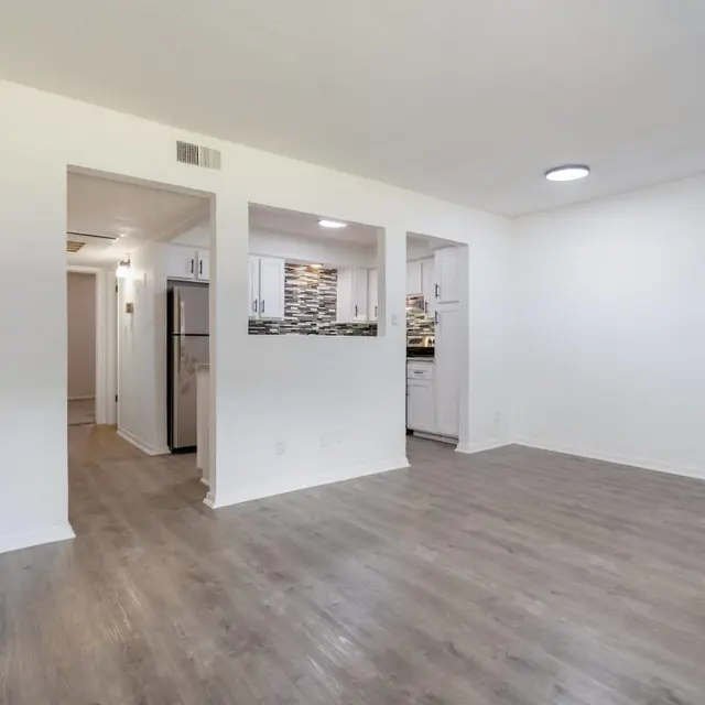 A spacious, bright living area of an apartment with light-colored walls and wood-style flooring. Two doorways lead to kitchen areas, one with modern cabinets and a backsplash visible. A ceiling light fixture illuminates the room.