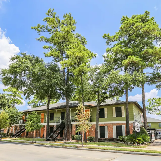 A green apartment complex surrounded by tall trees and a blue sky with fluffy clouds.