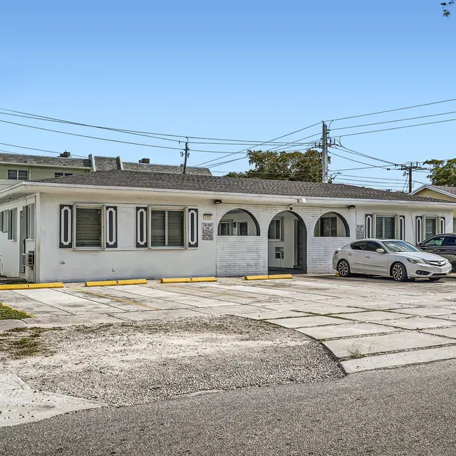 A small apartment complex view featuring a white building with arched entrances and parking spaces in front.