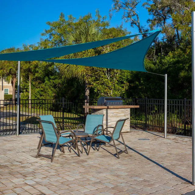 A shaded outdoor seating area featuring several chairs and a table, enclosed by a fence, with trees and a building in the background.