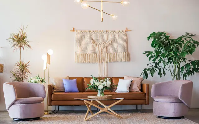 A stylish living room with a brown sofa, two purple chairs, a round coffee table, plants, and a decorative wall hanging.