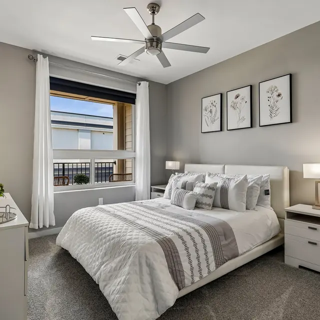 A modern bedroom featuring a neatly made bed with decorative pillows, two nightstands, and a large window with natural light. The walls are painted in a light gray tone, and there are framed botanical prints above the bed.