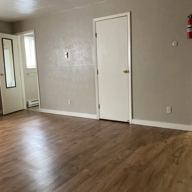 A spacious living room with light-colored walls and wooden laminate flooring. A door leads to another room, and there's an open doorway to a kitchen area in the background. An air conditioning unit is mounted on one wall, and a fire extinguisher hangs nearby.