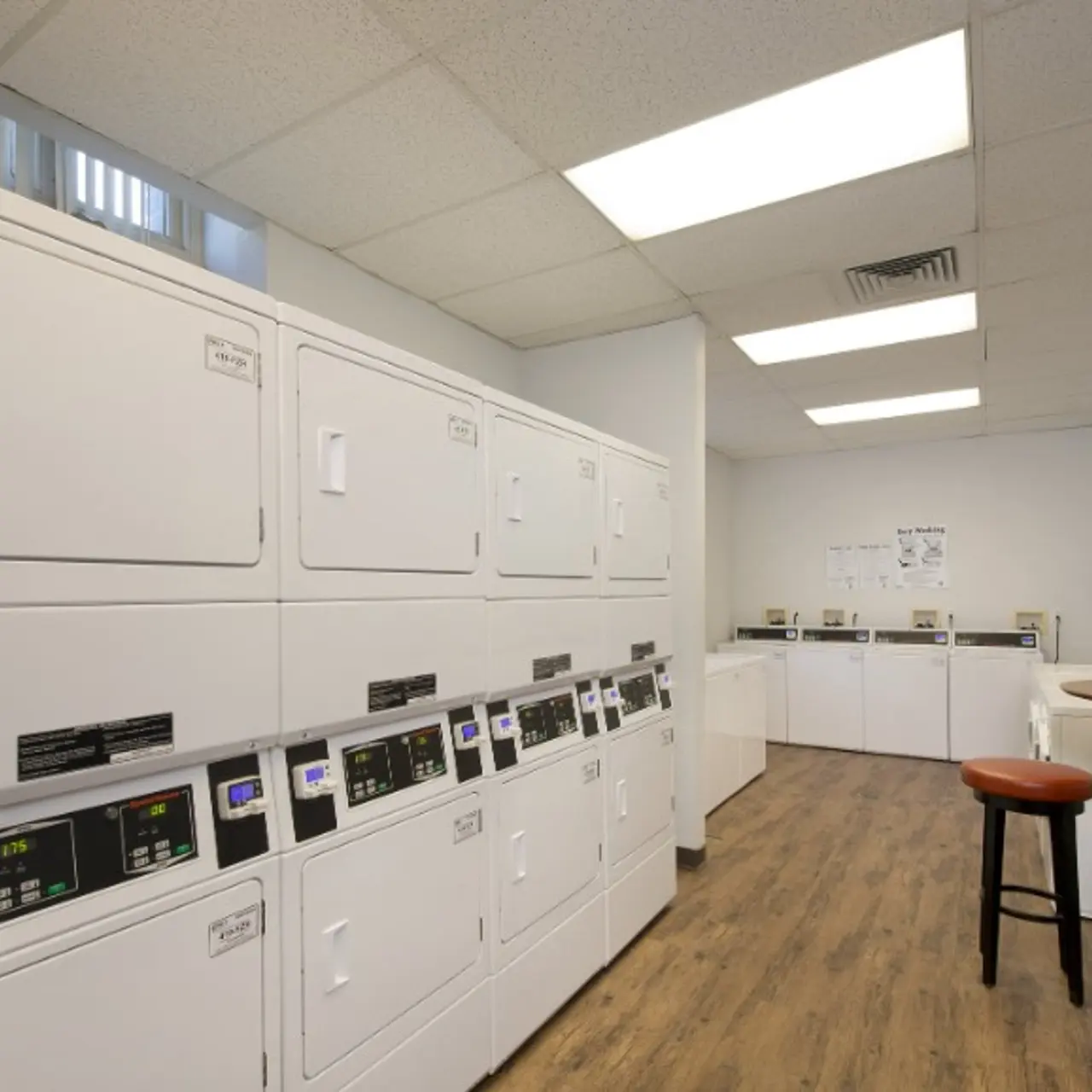 A modern laundry room with white washing machines and dryers lined against the wall, featuring a wooden floor and a seating area with two stools.