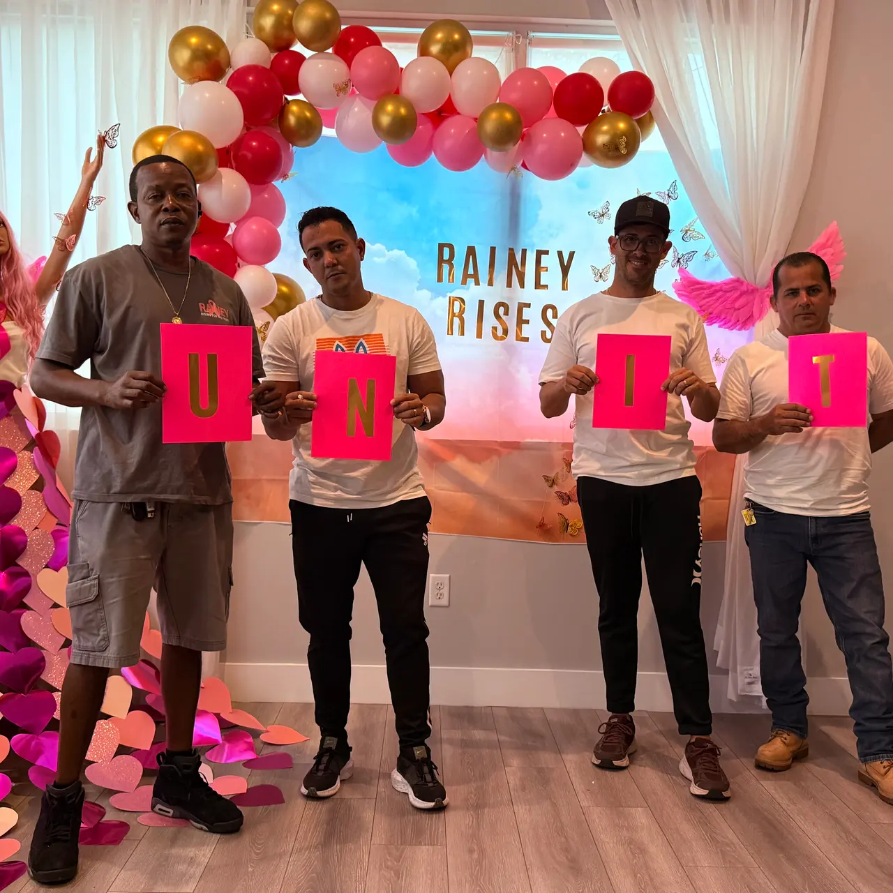 A group of five individuals holding colorful letters that spell 'UNITY' in front of a decorative backdrop with balloons and a figurative representation of a person with angelic wings.