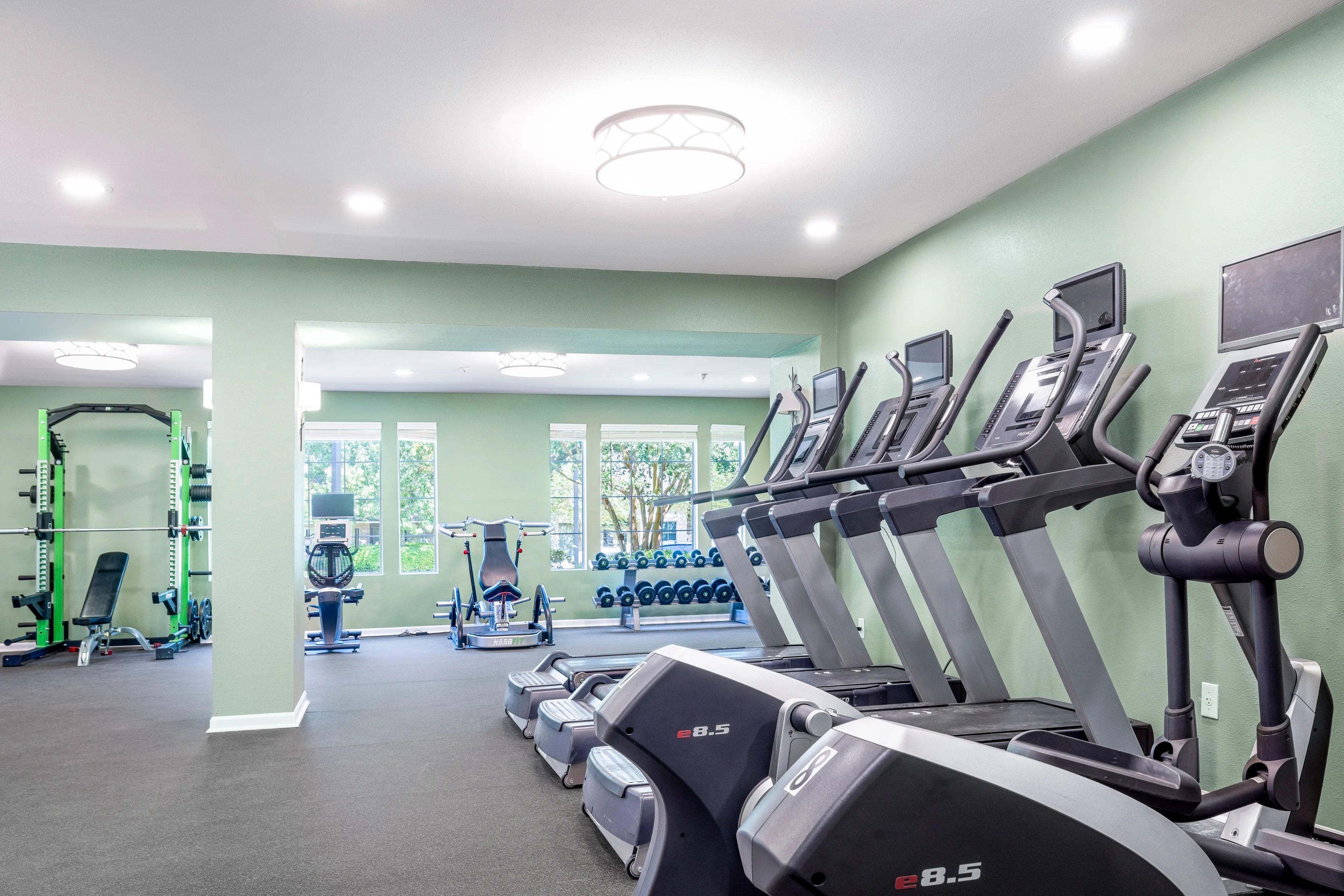 Fitness Center in Apartment Community An interior view of a fitness center with several elliptical machines and free weights, featuring a green wall and large windows letting in natural light.