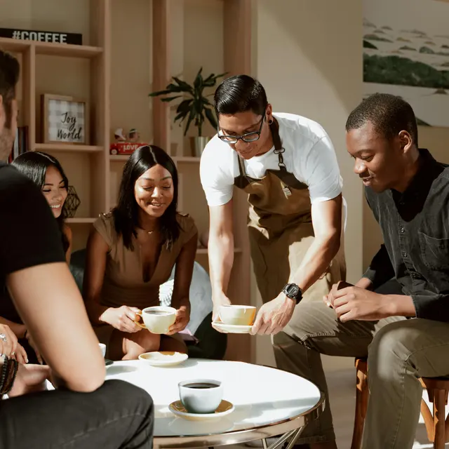Coffee Gathering Among Friends A group of five people in a bright, contemporary setting enjoying a coffee gathering. One person in an apron is serving coffee to the others seated around a low table, smiling and engaging with each other.