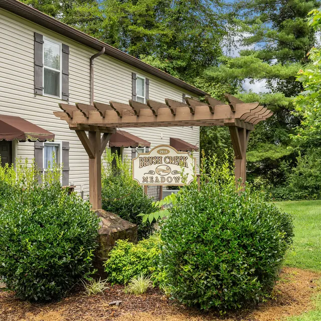 A two-story house with brown shutters and a decorative wooden trellis in front, surrounded by green shrubs and trees.