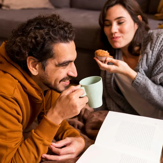 Cozy Couple Moments A couple enjoying a cozy moment at home, sitting on the floor wrapped in a blanket. The man is holding a light green mug and smiling at the woman, who is playfully holding a muffin toward him. They are surrounded by a warm and inviting home setting, with brown and gray decor.