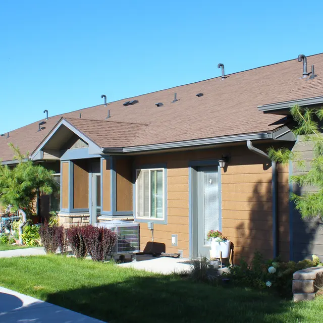 A row of residential houses with brown siding and pitched roofs. The houses are surrounded by greenery and gardens, with a well-maintained walkway in front.