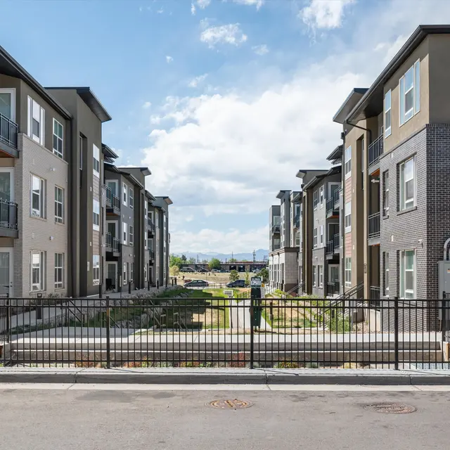 A view of a modern apartment complex with two rows of buildings on either side, featuring balconies and various architectural designs. A pathway runs between the buildings, leading to an open area in the center.