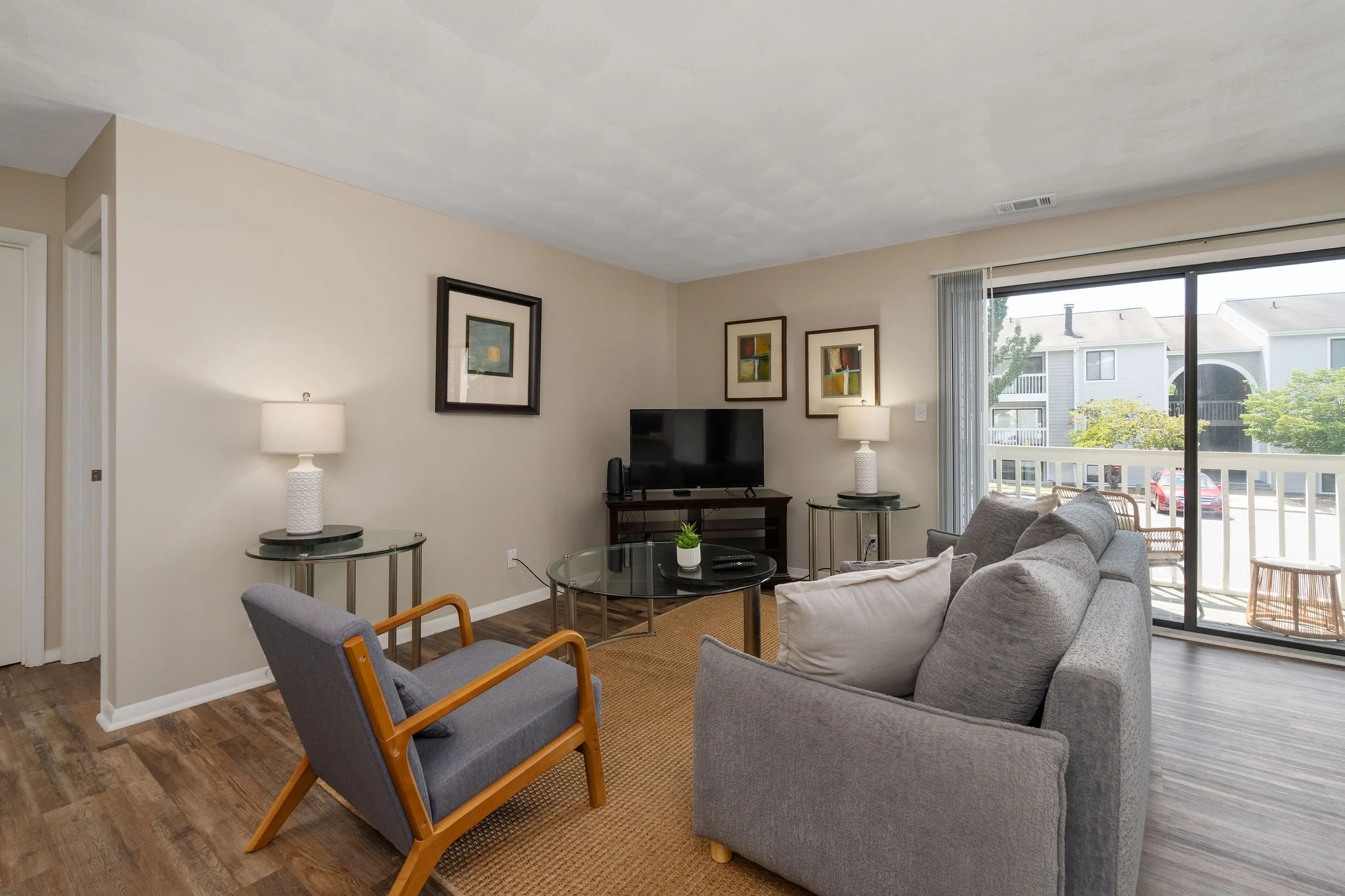 A cozy living room featuring a gray sofa, wooden chairs, and a television on a stand, with natural light coming through a large window.