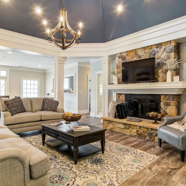 A stylish modern living room featuring a chandelier, a stone fireplace, a large TV mounted above it, and a comfortable seating arrangement with a coffee table. The room has a cozy carpet and elegant decor.