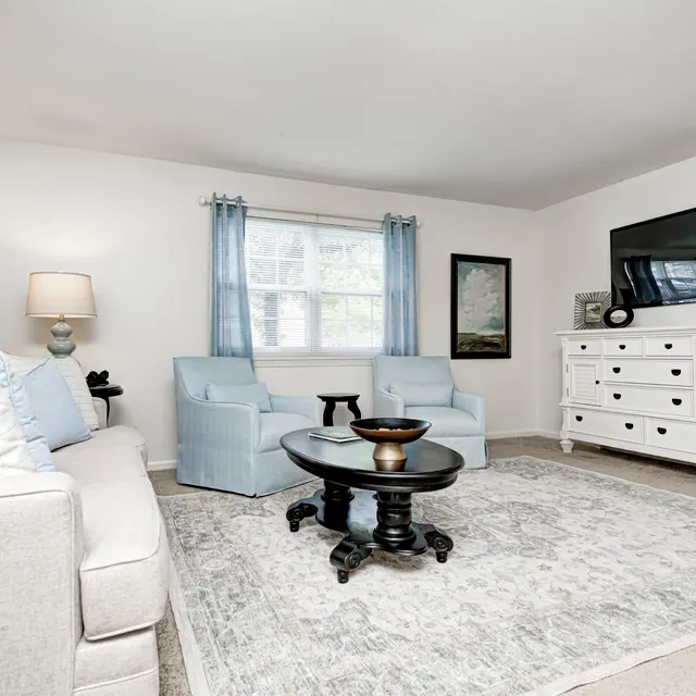 A cozy living room featuring a light-colored sofa, two blue armchairs, a round black coffee table, and a large dresser with a TV on top. There are light curtains on the windows and a soft area rug on the floor.