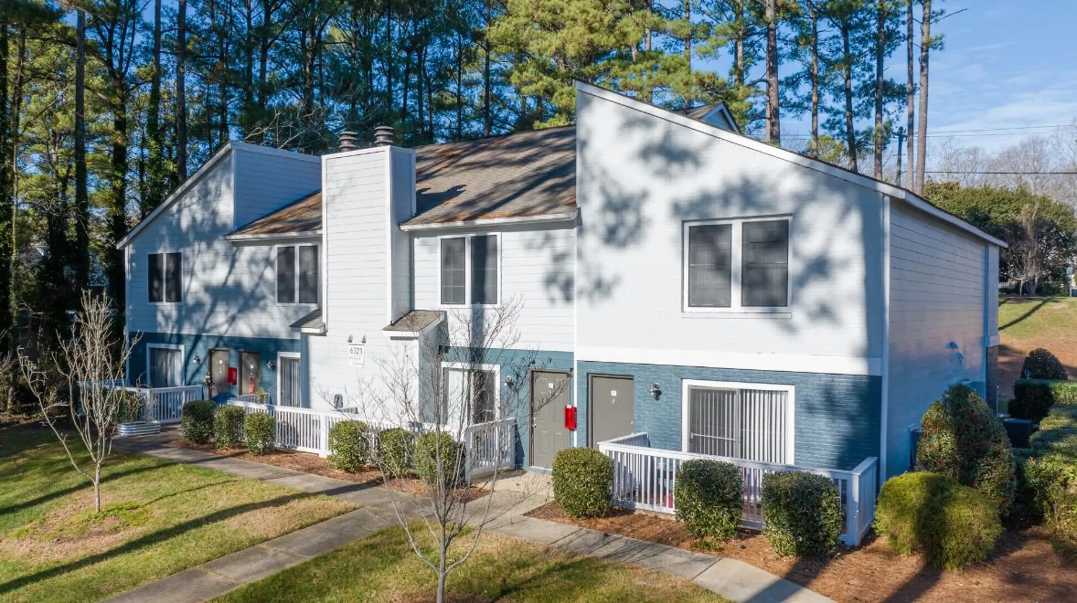 Aerial view of an apartment complex surrounded by trees. The buildings are two-story with white and light blue siding, featuring balconies and a neatly landscaped lawn with shrubs.