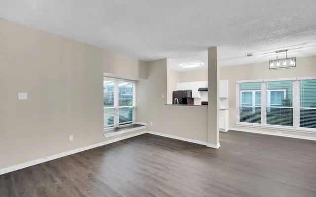A spacious and modern living room with a wooden floor, featuring large windows and an open kitchen area in the background.