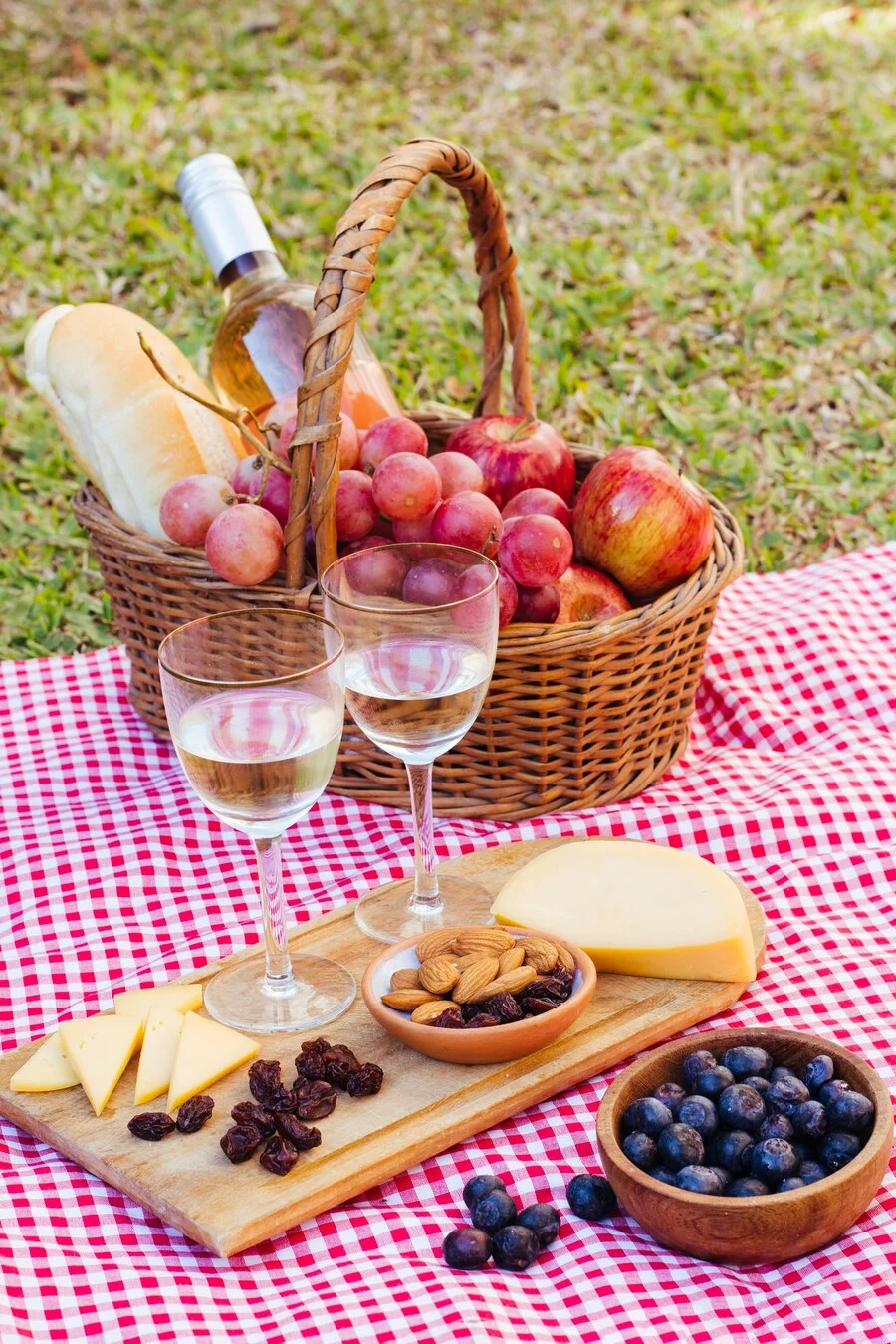 A picnic setup featuring a basket filled with fruit, a bottle of wine, two glasses of white wine, a wooden board with cheese, nuts, and raisins, and a bowl of blueberries, all on a checkered blanket in a grassy area.