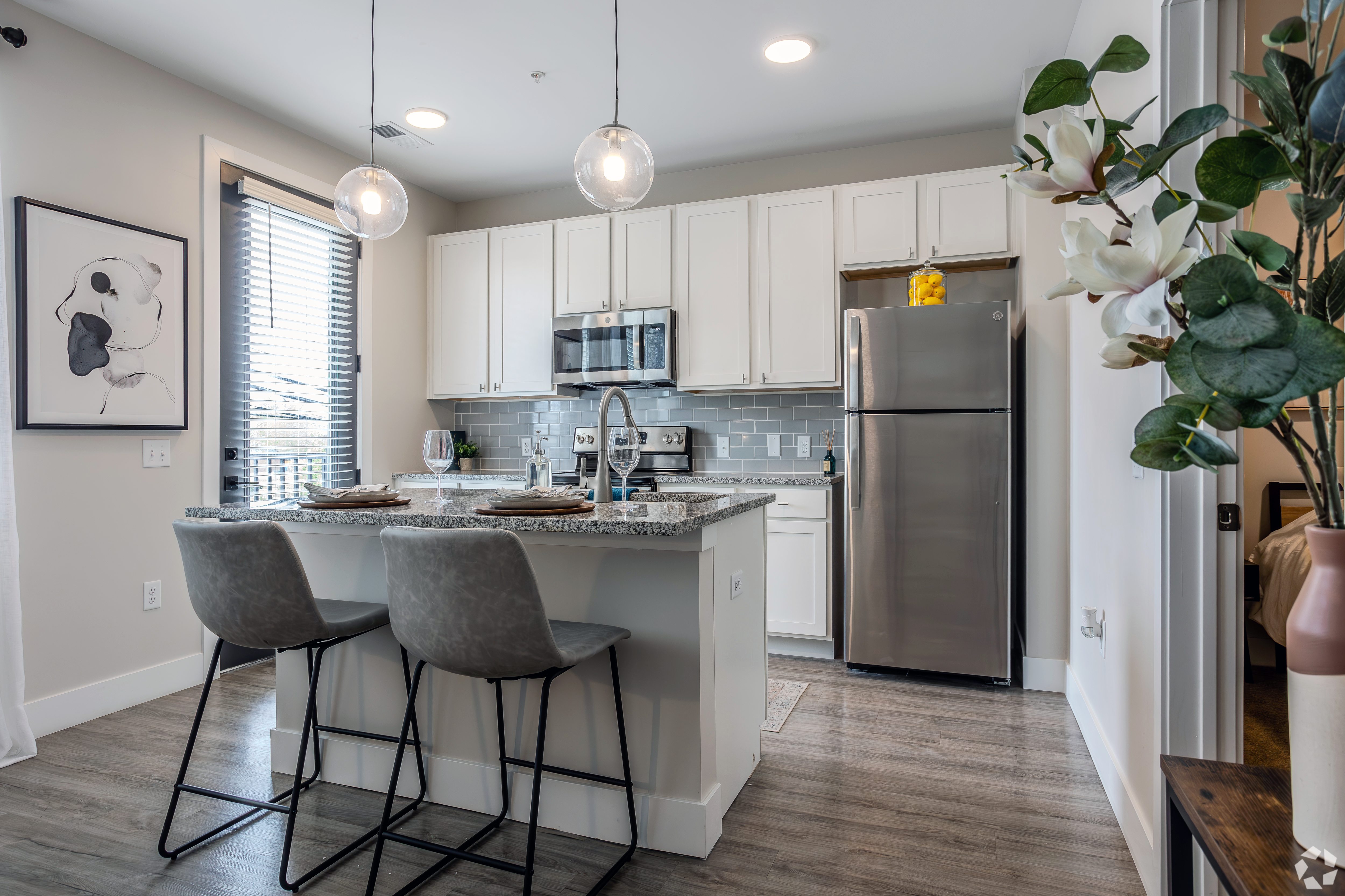 A modern kitchen featuring a large island, bar stools, stainless steel appliances, and a window with blinds. Decorative plants are in the foreground.