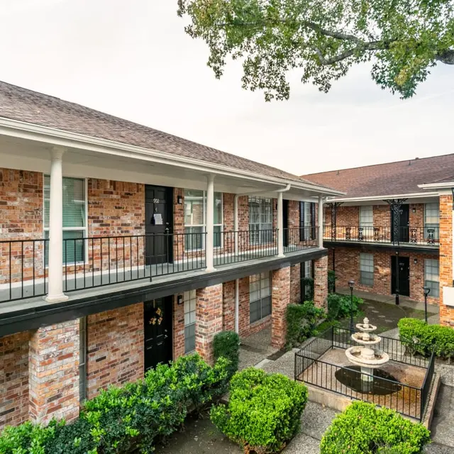 Exterior view of an apartment complex with brick walls, balconies, and a central courtyard featuring a fountain and landscaping.