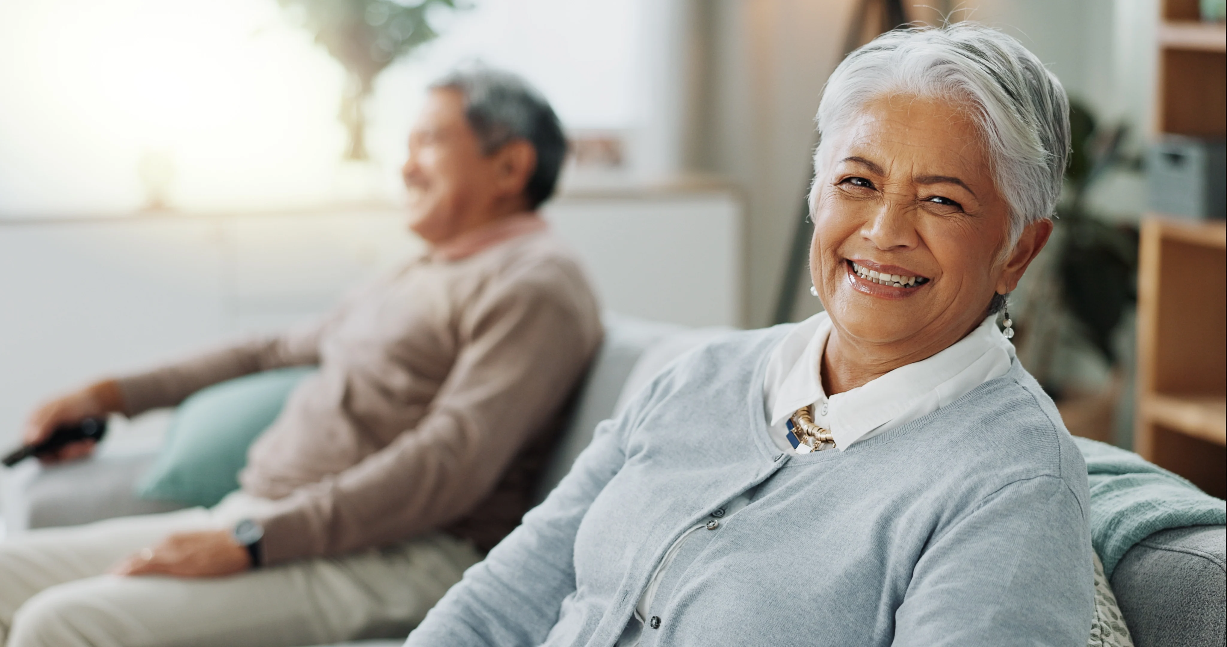 A joyful elderly woman smiling at the camera while sitting on a couch. In the background, an elderly man is lounging with a remote control, appearing relaxed.