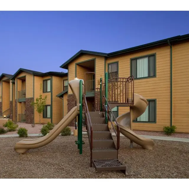 Children's Slide at Apartment Complex A view of an apartment complex with a beige and green color scheme. There is a metal staircase leading up to a walkway with a spiral slide at the side. The setting is surrounded by gravel and a few small plants.
