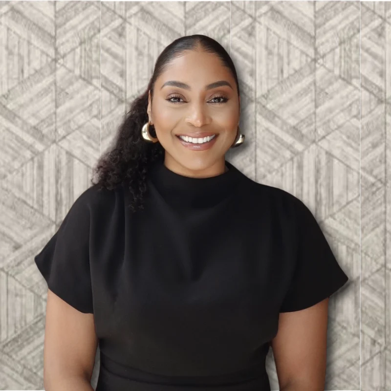 Smiling Woman in Black Top A woman with long dark hair and a smile, wearing a black top, standing in front of a patterned wall.