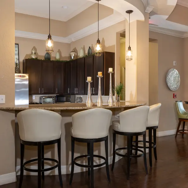 A modern kitchen bar area featuring four white upholstered stools, dark wood cabinets, and pendant lighting, with a small dining area in the background.