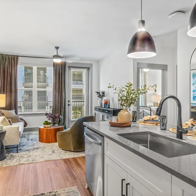 A modern apartment interior featuring a kitchen view into a cozy living area. The kitchen has white cabinetry and a dark countertop. Sunlight filters through large windows, illuminating the comfortable seating area with two armchairs and a sofa. A round coffee table sits on a light-colored rug, and decorative items adorn the space, enhancing its inviting atmosphere.